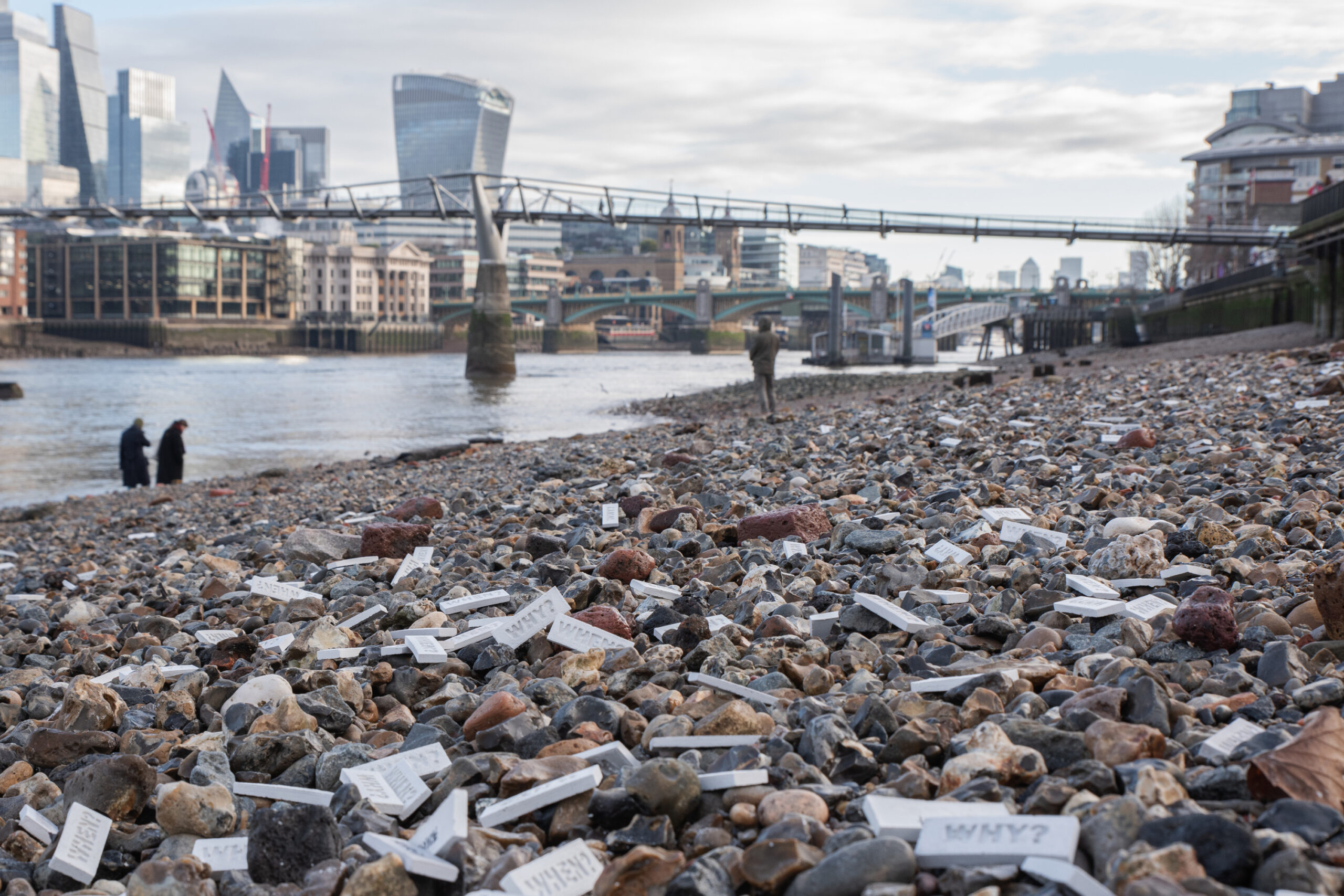 Concrete “WHEN?” and “WHY?” tiles scattered across the Thames foreshore at low tide, with bridges and the City skyline in the background.